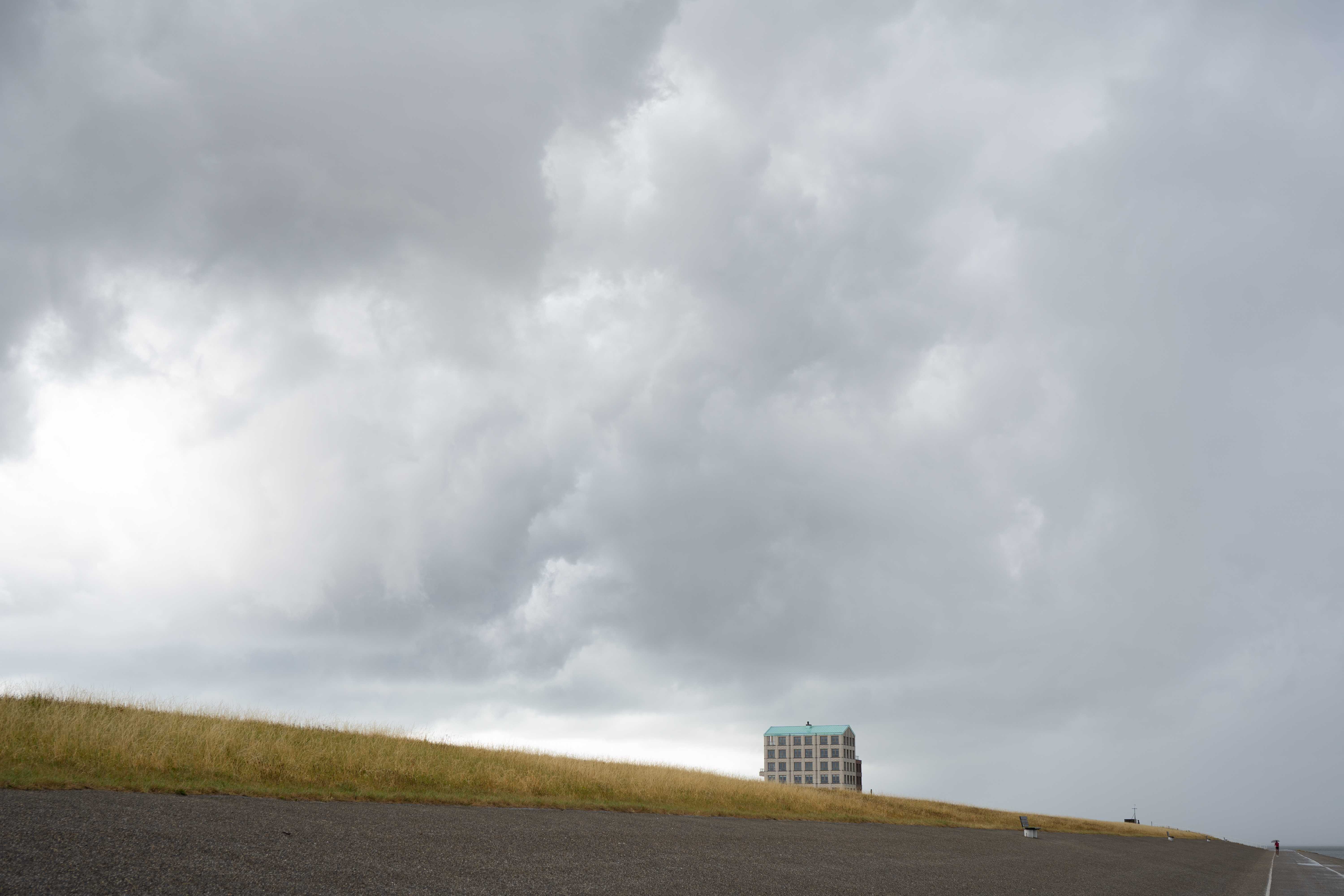 A photo showing an abstract image of a sea dike covered in asphalt and grass, and a cloudy sky above. A single building is visible sticking out above the dike.